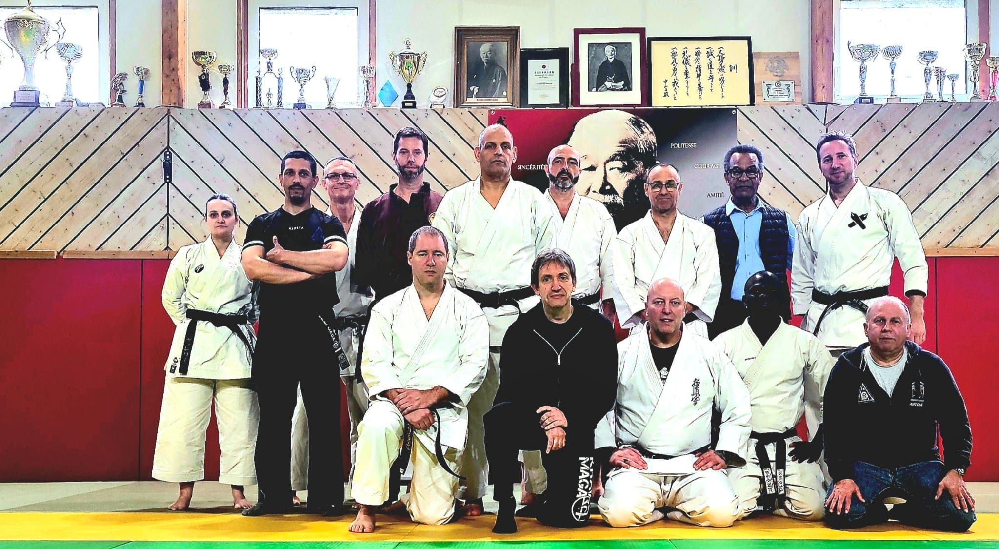 Pratiquants en kimono réunis dans un dojo, devant des portraits accrochés au mur.